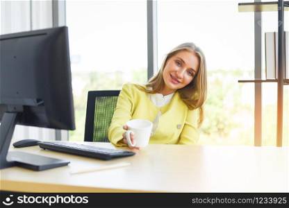 Smiling woman sitting at her desk in office and holding a coffee cup / Happy business woman sitting in office , businesswoman working place