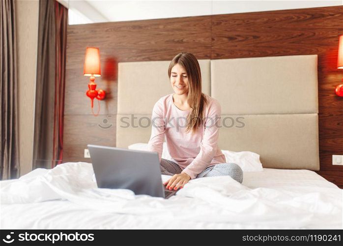 Smiling woman sitting in bed and uses laptop, good morning, bedroom interoir on background. Woman sitting in bed and uses laptop, good morning