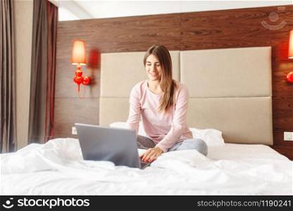 Smiling woman sitting in bed and uses laptop, good morning, bedroom interoir on background. Woman sitting in bed and uses laptop, good morning