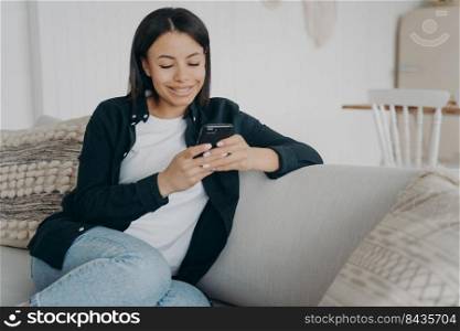 Smiling woman sitting on couch with smartphone. Young female holding phone, share news messages with friend in social networks, enjoy internet services, shopping online at home.. Smiling woman sitting on couch with smartphone chatting in social networks, shopping online at home
