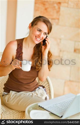 Smiling woman sitting on terrace with laptop and cup of tea and speaking mobile &#xA;