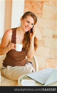 Smiling woman sitting on terrace with laptop and cup of tea and speaking mobile &#xA;