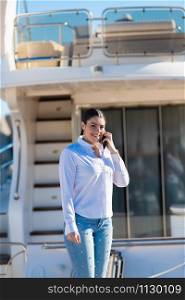 Smiling woman standing next to a boat looks at the camera while talking on her phone. Leisure concept.. Woman smiles standing next to a boat