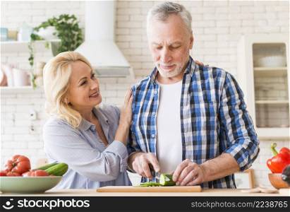 smiling woman supporting her husband cutting cucumber with knife table kitchen