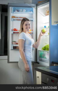 Smiling woman taking green apple from fridge at night