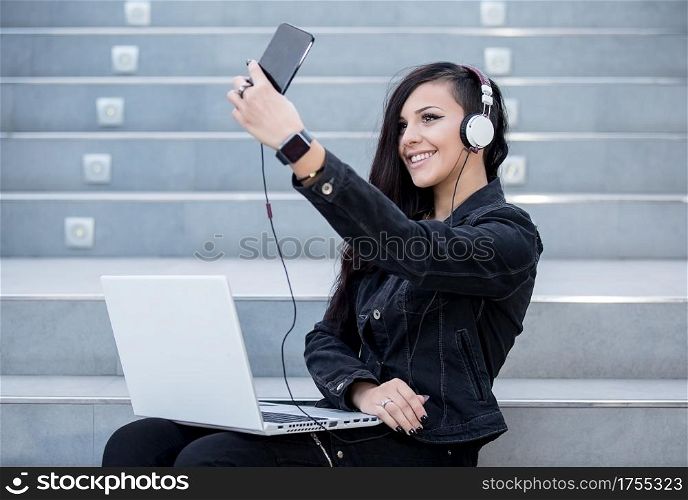 Smiling woman taking selfie?while listening to music and using laptop on stairs.. Woman with gadgets on steps