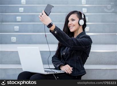 Smiling woman taking selfie?while listening to music and using laptop on stairs.. Woman with gadgets on steps