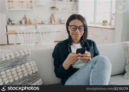 Smiling woman uses smartphone chatting in social networks, shopping in online store, sitting on couch at home. Female answering messages on phone, browsing the Internet. E-commerce, gadget addiction.. Smiling woman uses smartphone chatting in social networks, shopping online, sitting on couch at home