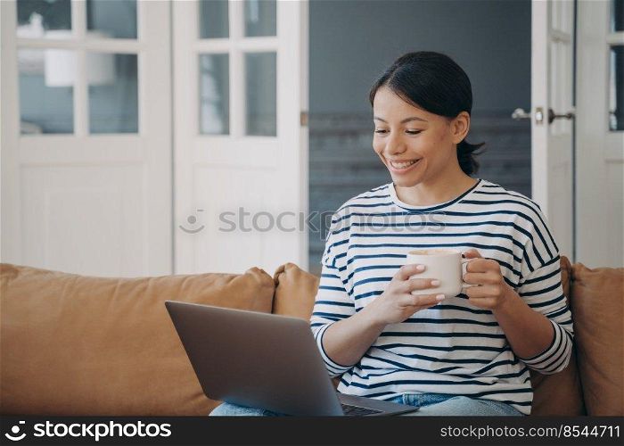 Smiling woman using laptop relaxing on comfortable sofa at home, happy girl holding cup of coffee, watching movies, shopping online on computer, communicating by video call, spending lazy weekends. . Smiling girl with coffee cup watching movie or communicating by video call at laptop on sofa at home
