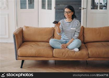 Smiling woman watching television series or movie, changes TV channel by remote control, sitting on modern sofa in living room at home. Happy female wearing glasses rests choosing film to see.. Smiling woman watching television series or movie changes TV channel, sitting on modern sofa at home
