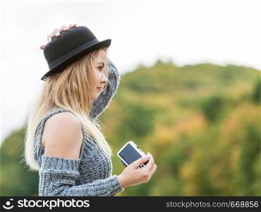 Smiling woman wearing black hat and grey sweater taking pictures using digital camera on outdoor photo session. Female having passion.. Woman in hat taking outdoor pictures