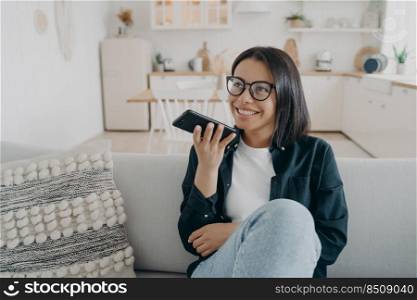 Smiling woman wearing glasses hold smartphone, chatting, sitting on couch at home. Female use virtual assistant on phone, talks with friend using speaker phone, records voice message in social network. Female holds smartphone, talks using speaker phone, records voice message, sitting on sofa at home