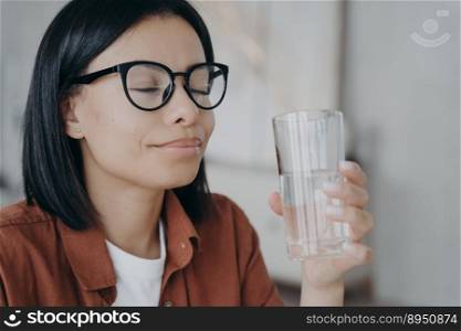 Smiling woman wearing glasses holds a glass of water. Calm happy female with closed eyes enjoy pure clear mineral water, refresh the body, preventing dehydration. Healthy lifestyle habbit, wellness.. Happy woman wearing glasses holds glass of pure water, closed eyes. Healthy lifestyle, wellness