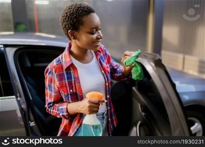 Smiling woman wipes a car with a rag, hand auto wash station. Car-wash industry or business. Female person cleans her vehicle from dirt outdoors. Smiling woman wipes car with rag, hand auto wash