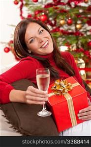 Smiling woman with Christmas present and glass of champagne in front of tree