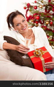 Smiling woman with Christmas present and glass of champagne in front of tree