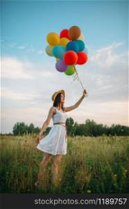 Smiling woman with colorful balloons walking in green field. Pretty girl on summer meadow at sunny day. Smiling woman with balloons walking in green field
