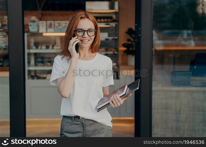 Smiling woman with laptop and notebook in hand enjoying talk on cellphone, consulting client or customer while standing near coffee shop, happy young redhead businesswoman talking smartphone outdoor. Smiling woman with laptop and notebook in hand enjoying talk on cellphone near coffee shop