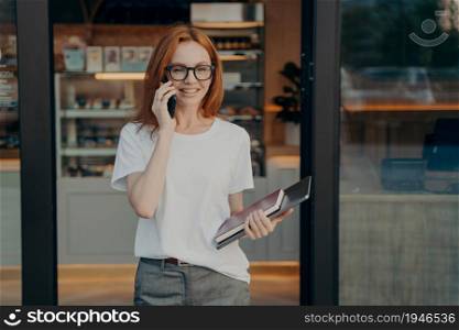 Smiling woman with laptop and notebook in hand enjoying talk on cellphone, consulting client or customer while standing near coffee shop, happy young redhead businesswoman talking smartphone outdoor. Smiling woman with laptop and notebook in hand enjoying talk on cellphone near coffee shop