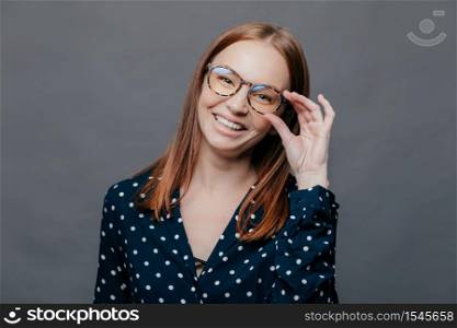 Smiling woman with pleased facial expression, keeps hand on rim of spectacles, wears black polka dot blouse, rejoices promotion at work, poses over grey background. People, happiness concept