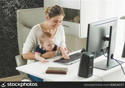 Smiling woman working at home and holding her baby son on lap