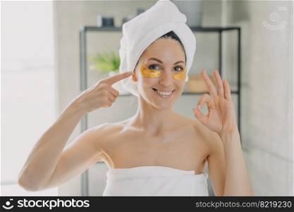 Smiling woman wrapped in bath towels recommending eye patches, showing ok gesture in bathroom. Satisfied pretty female advertising hydrogel undereye patch for dark circles bags treatment. Skin care.. Pretty woman advertising anti wrinkles anti age eye patches showing ok gesture in bathroom. Skincare