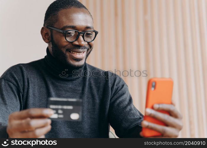 Smiling young African American businessman using credit card and smartphone while sitting at workplace in modern office.Cheerful employee doing online purchase or contacts with banking support service. Smiling African American businessman using credit card and smartphone while sitting at workplace