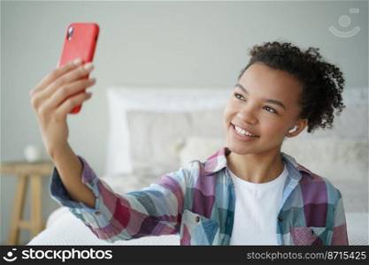 Smiling young african american girl wearing earphone, holding smartphone, chatting online by video call at home. Happy teenager lady blogger takes pictures, makes selfie photography in bedroom.. Smiling young african american girl in earphone holding phone, chatting online by video call at home