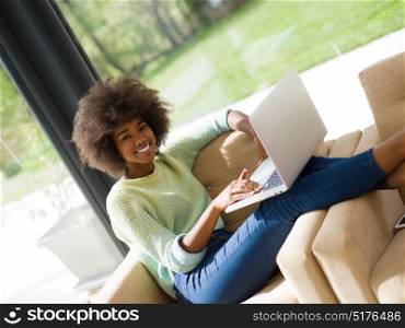 Smiling young African American woman using laptop while relaxing on sofa at luxury home