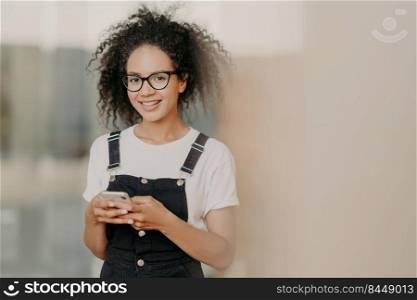 Smiling young Afro female holds modern cell phone, uses mobile app, lives in world of modern technologis, addicted to social networks, chats with friends online, dressed in fashionable outfit