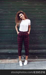 Smiling young arabic woman with black curly hairstyle. Arab girl in casual clothes in the street. Happy female wearing white t-shirt and checked pants against urban blinds.