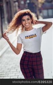 Smiling young arabic woman with black curly hairstyle. Arab girl in casual clothes in the street. Happy female wearing white t-shirt and checked pants.