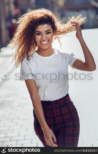 Smiling young arabic woman with black curly hairstyle. Arab girl in casual clothes in the street. Happy female wearing white t-shirt and checked pants.