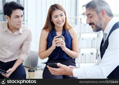Smiling young Asian and Senior Caucasian businesspeople teamwork relaxing and brainstormimg with tablet for project work and businesswoman holding a mug of hot coffee at office. Focus asian woman.