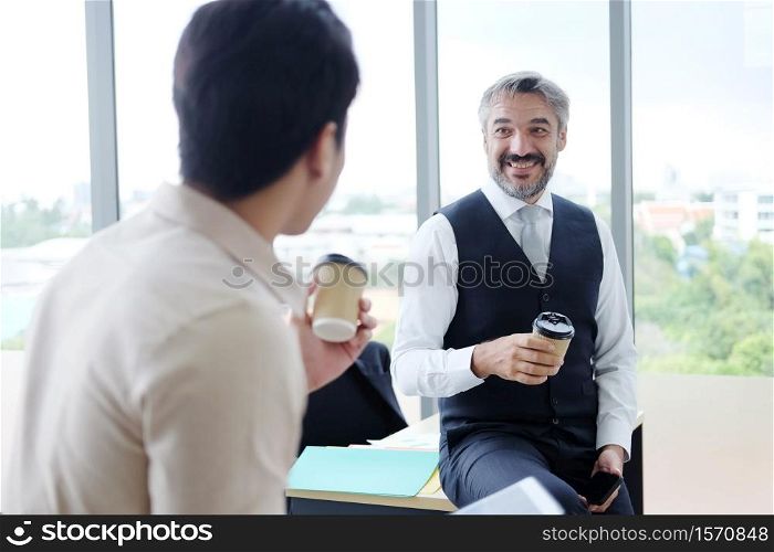 Smiling young Asian and Senior Caucasian businesspeople teamwork relaxing and drinking a mug of hot coffee together at modern office. Business and lifestyle concept.