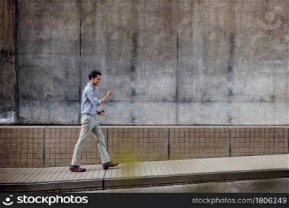 Smiling Young Asian Businessman in Casual wear Using Mobile Phone while Walking by the Urban Building Wall. Lifestyle of Modern People. Side View. Full Length