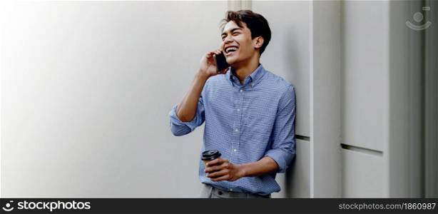 Smiling Young Asian Businessman Using Mobile Phone in the City. Standing with Coffee by the Wall. more copy space with Wide size