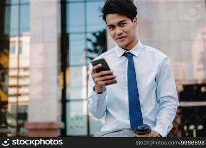Smiling Young Asian Businessman Using Mobile Phone in the City. Looking at the Camera