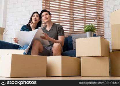 Smiling young Asian happy couple hold blueprint for home decoration ideas at moving day in their new home after buying real estate. Concept of starting a new life for a newly married couple.