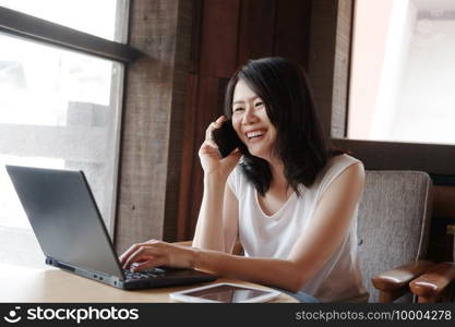 Smiling young Asian woman is calling with smartphone and online working with laptop in living room. Work from home for Covod-19 outbreak concept.