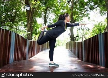 Smiling young Asian woman standing on wooden bridge for exercising and stretching the legs with Yoga for relax and meditation body after running in the garden. Sport and healthy Concept