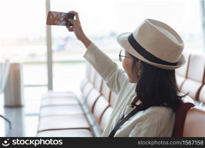Smiling young Asian woman traveler taking a selfie with mobile phone or smartphone while travel in vacation at international airport.