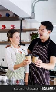 Smiling young barista couple loves Asian man and caucasian woman is hugging and holding cups of hot coffee together. Start up Coffee shop and cafe business concept.