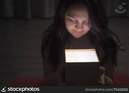 Smiling young brunette woman sitting on the floor and looking into the opened box from her hands, from which a light comes out.