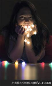 Smiling young brunette woman sitting on the floor in a dark room, holding lit string lights, enjoying their reflection and the atmosphere created.