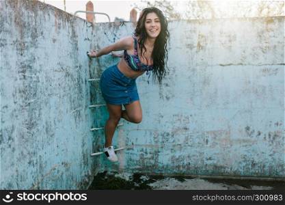 Smiling young brunette woman with bikini in an old empty pool playing with ladder