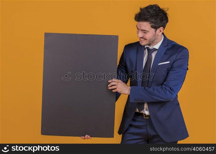 smiling young businessman holding blank black placard against orange backdrop