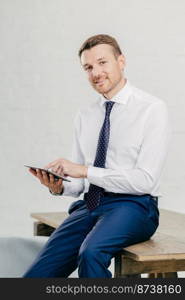 Smiling young businessman in elegant clothes happy to recieve income message on modern tablet computer, sits at wooden office desk in his cabinet, poses against white wall. Business concept.