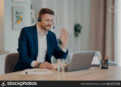 Smiling young businessman in headset and suit saying hello, waving to somebody and smiling while working remotely at home, having video call with colleagues or taking part in web conference. Happy businessman in headset having video call while working from home