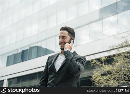 smiling young businessman standing front office building talking mobile phone